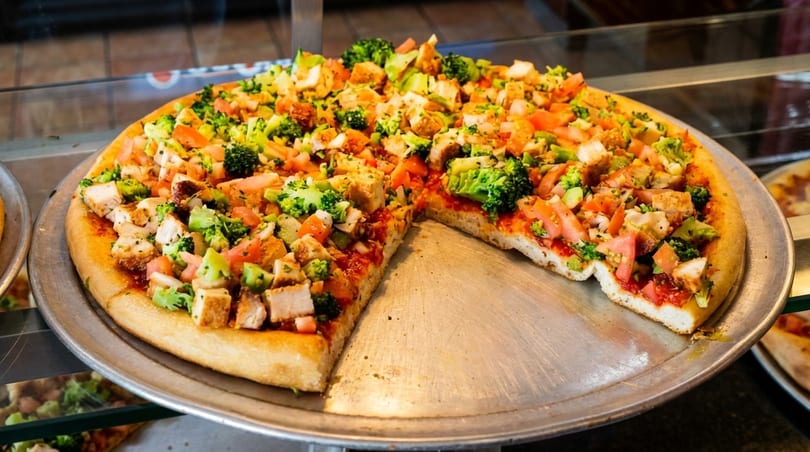 A large pizza with chicken, broccoli, and tomatoes, with one slice removed from the pan.