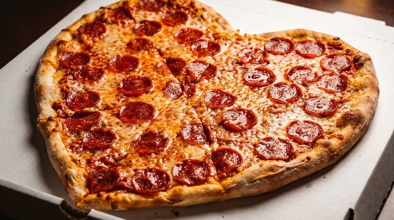 Heart-shaped pepperoni pizza with melted cheese, sliced and ready to eat in a white pizza box.
