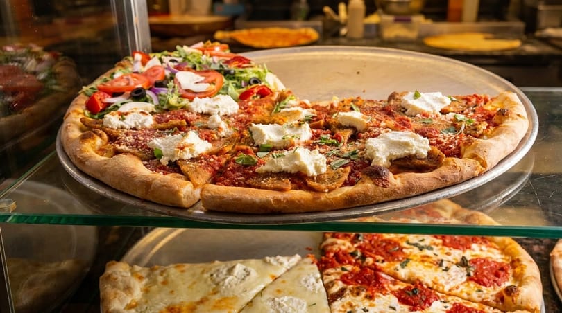 Two large pizzas, one with ricotta and tomato sauce, the other with salad toppings, in a display case.